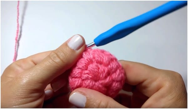 a person is knitting a pink crochet ball with a blue tool.