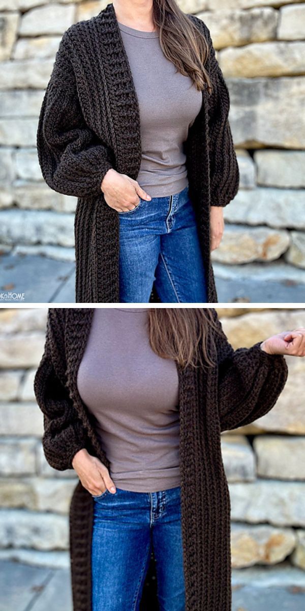 A woman wearing a long, brown crocheted cardigan paired with blue jeans and a grey top, standing against a stone wall background.