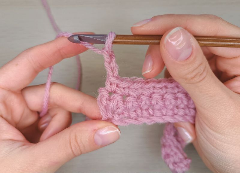 A piece of pink crocheted fabric in hands presenting how to do a half double crochet stitch