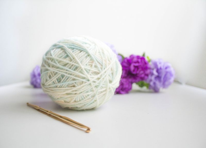 A ball of yarn with crochet hook and purple flowers in the background. The start tools before making a slip knot crochet