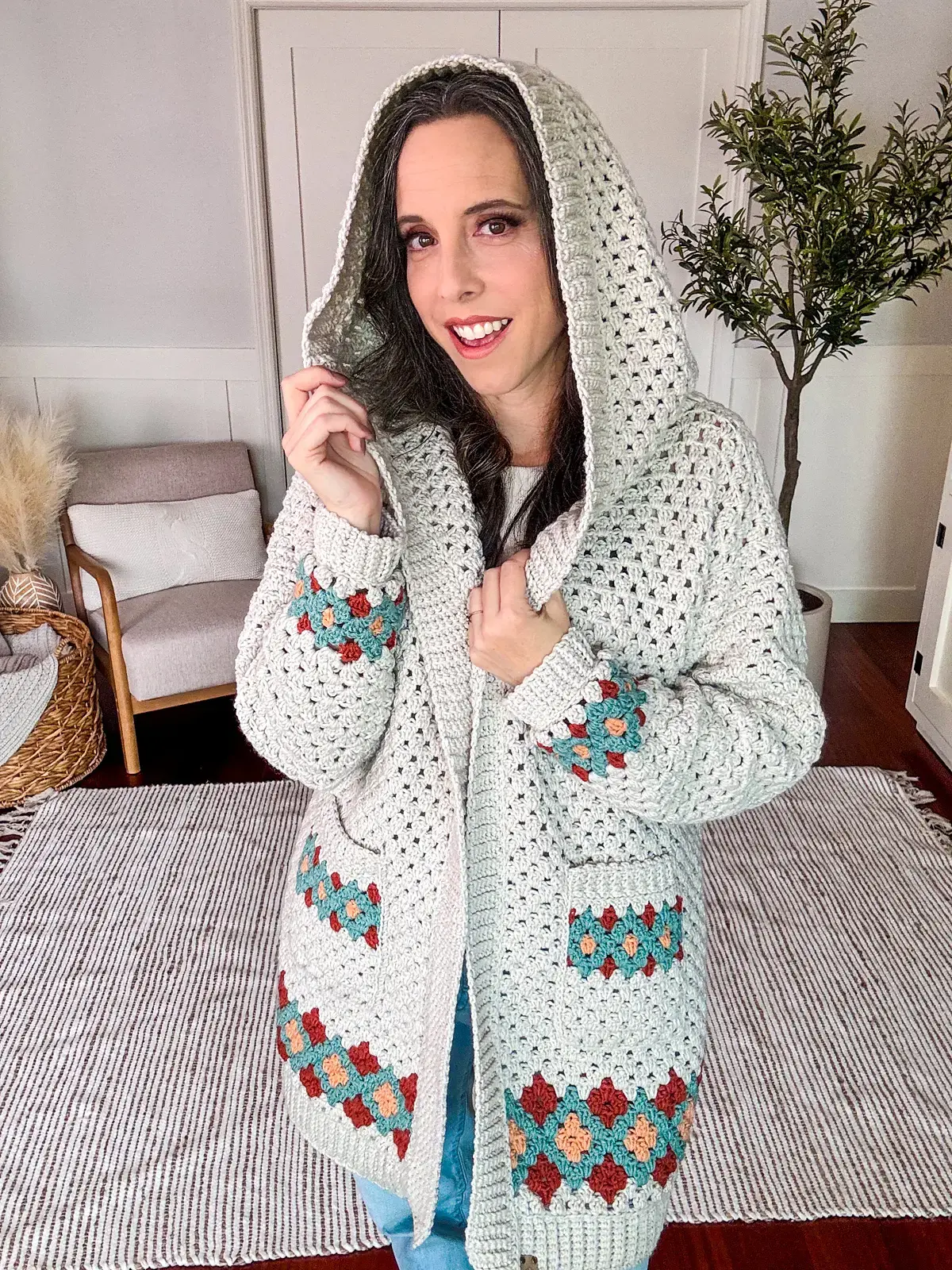 A person wearing a hooded, crocheted granny square cardigan with colorful patterns stands indoors, smiling.