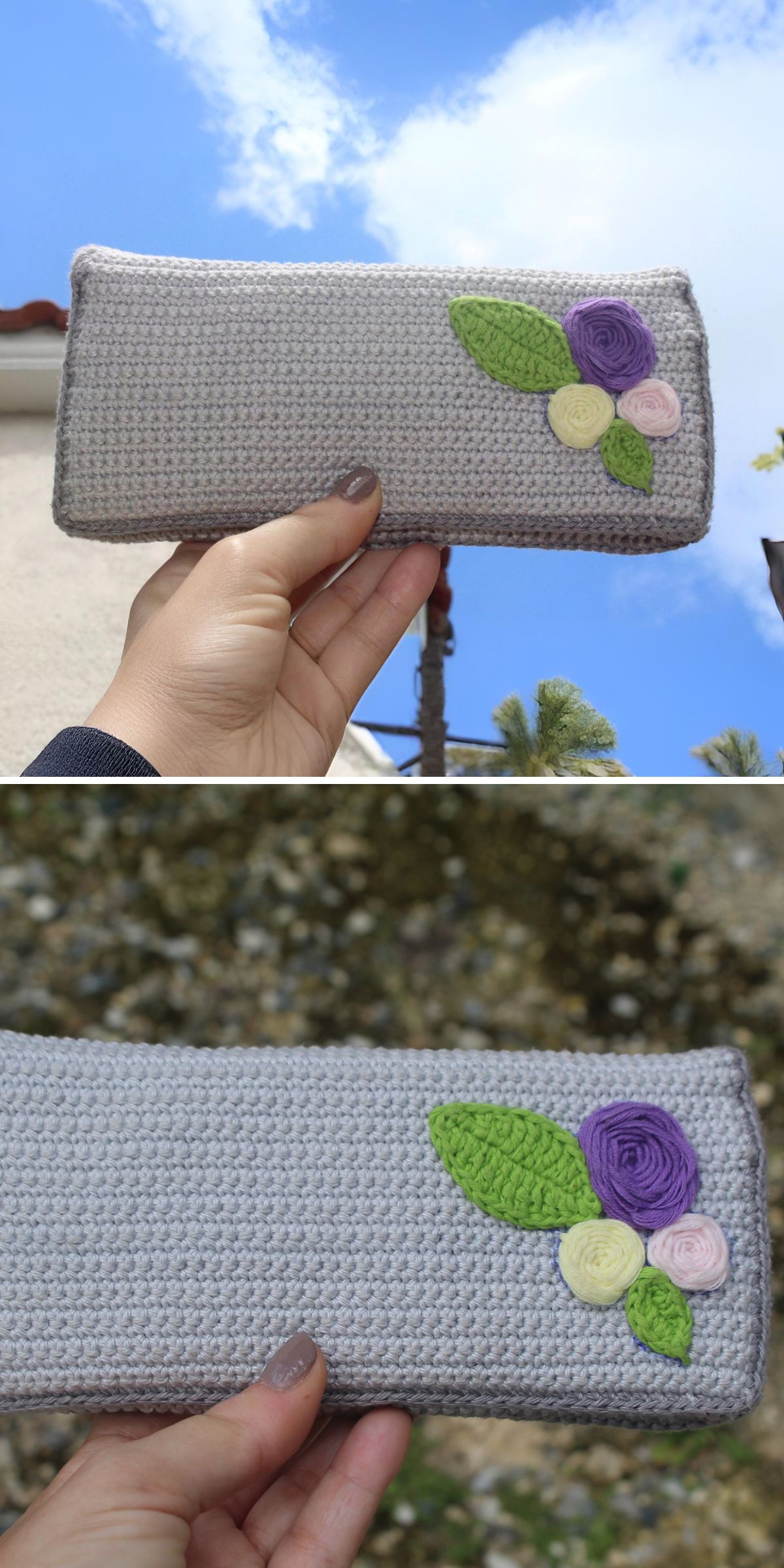 A hand holds a gray crochet clutch wallet adorned with two green leaves and three small flower appliqués in purple, yellow, and pink.