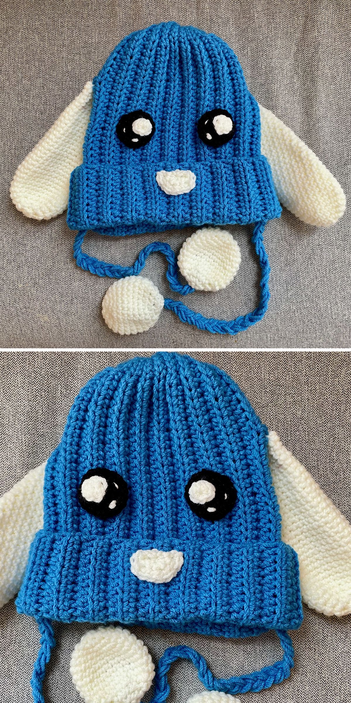 A blue crochet baby hat with large white eyes, a small white mouth, long cream-colored ears, and two pom-poms attached by braided strings, displayed on a gray surface.