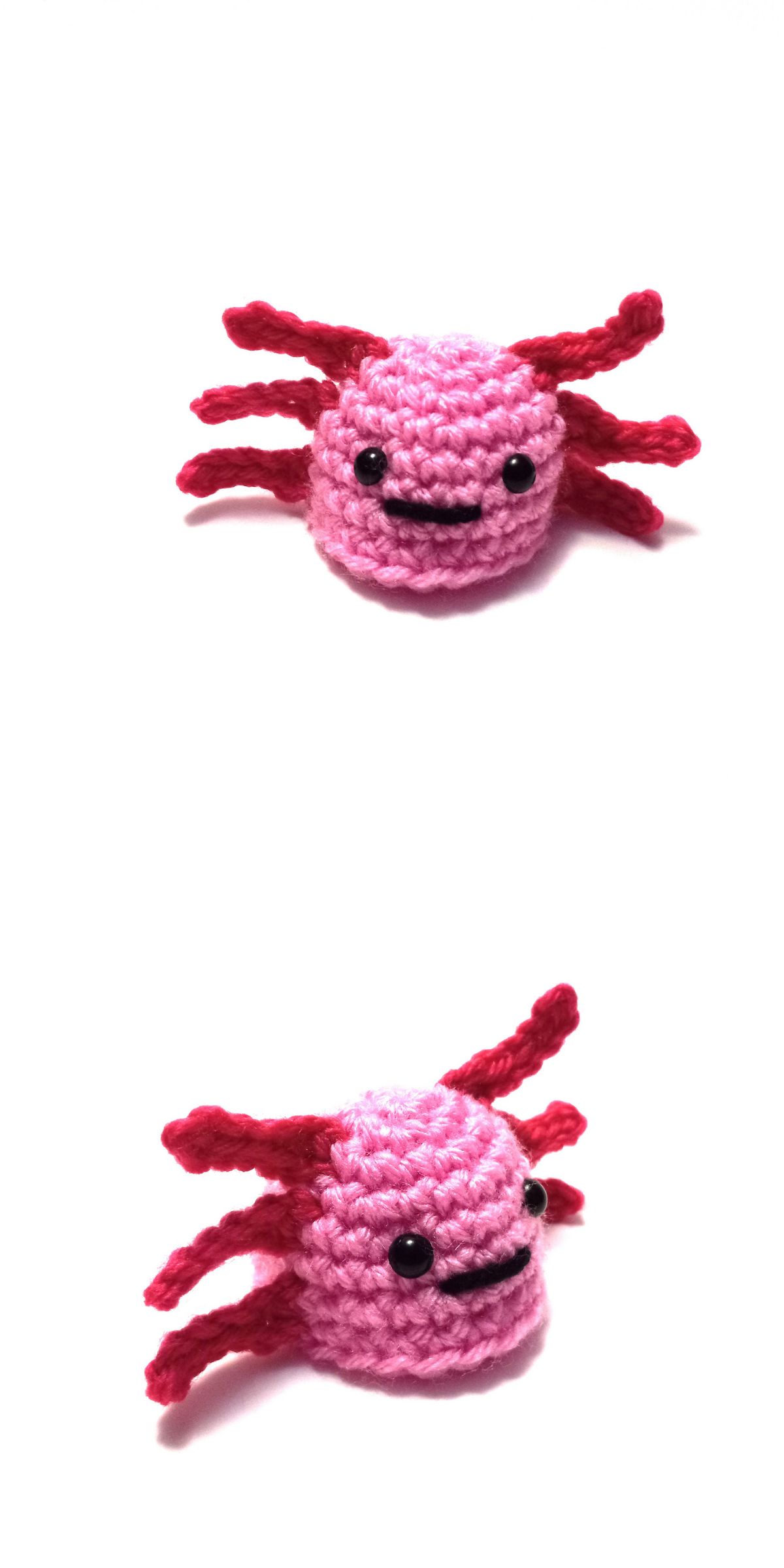 A small, crochet axolotl gat in pink and red with a smiling face is shown against a white background from two angles.
