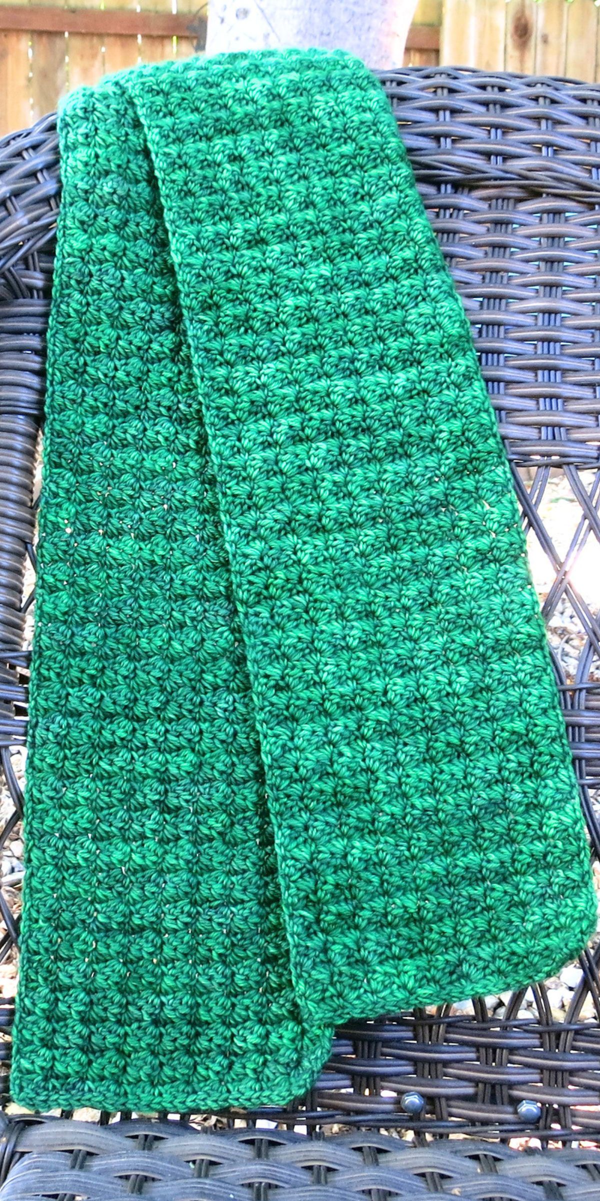 A green crocheted scarf, inspired by free crochet patterns in green, is draped over the back of a woven wicker chair outdoors.