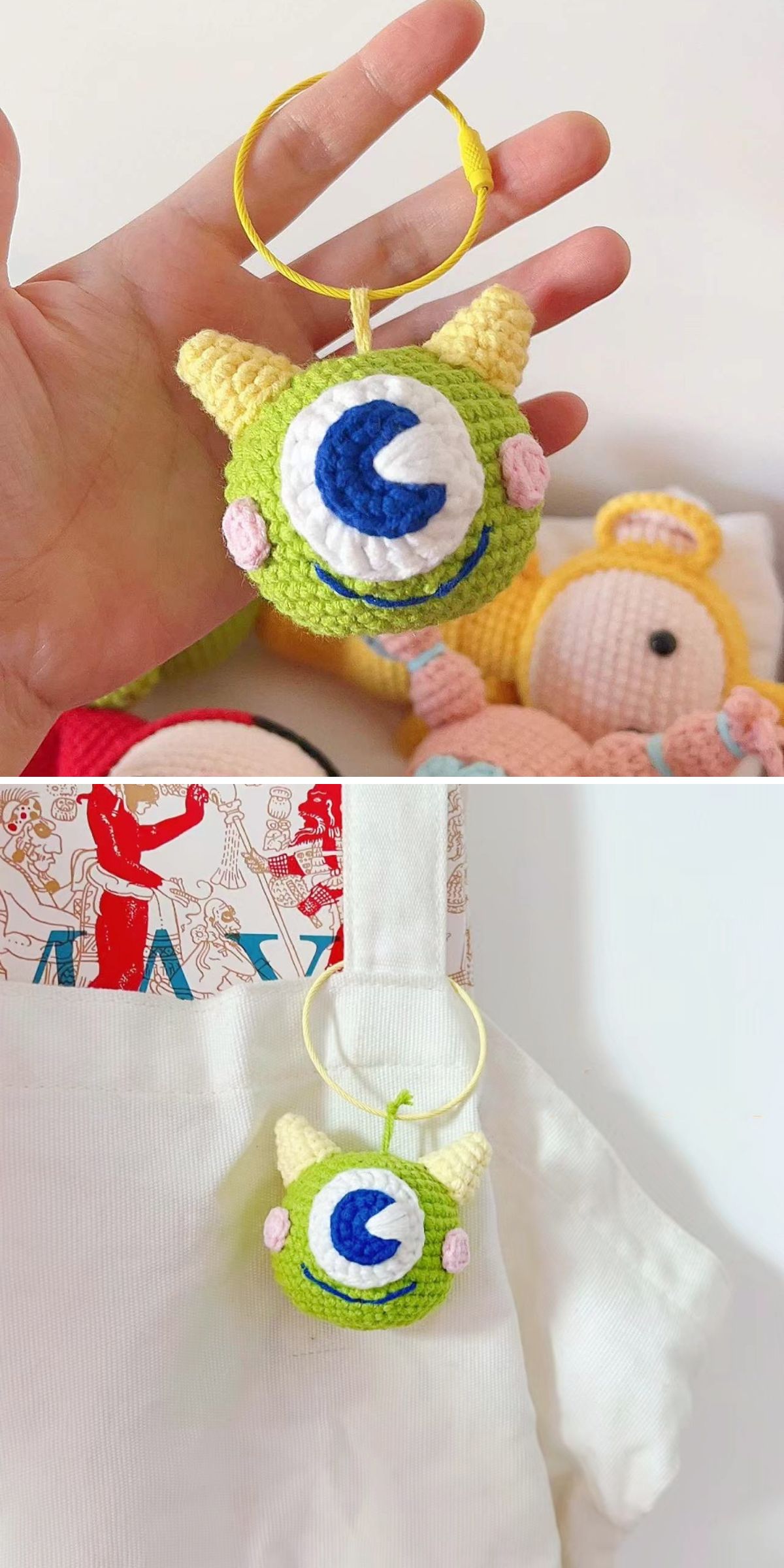 A crochet keychain shaped like a green monster with one large blue eye and yellow horns is shown in a hand and attached to a white tote bag.