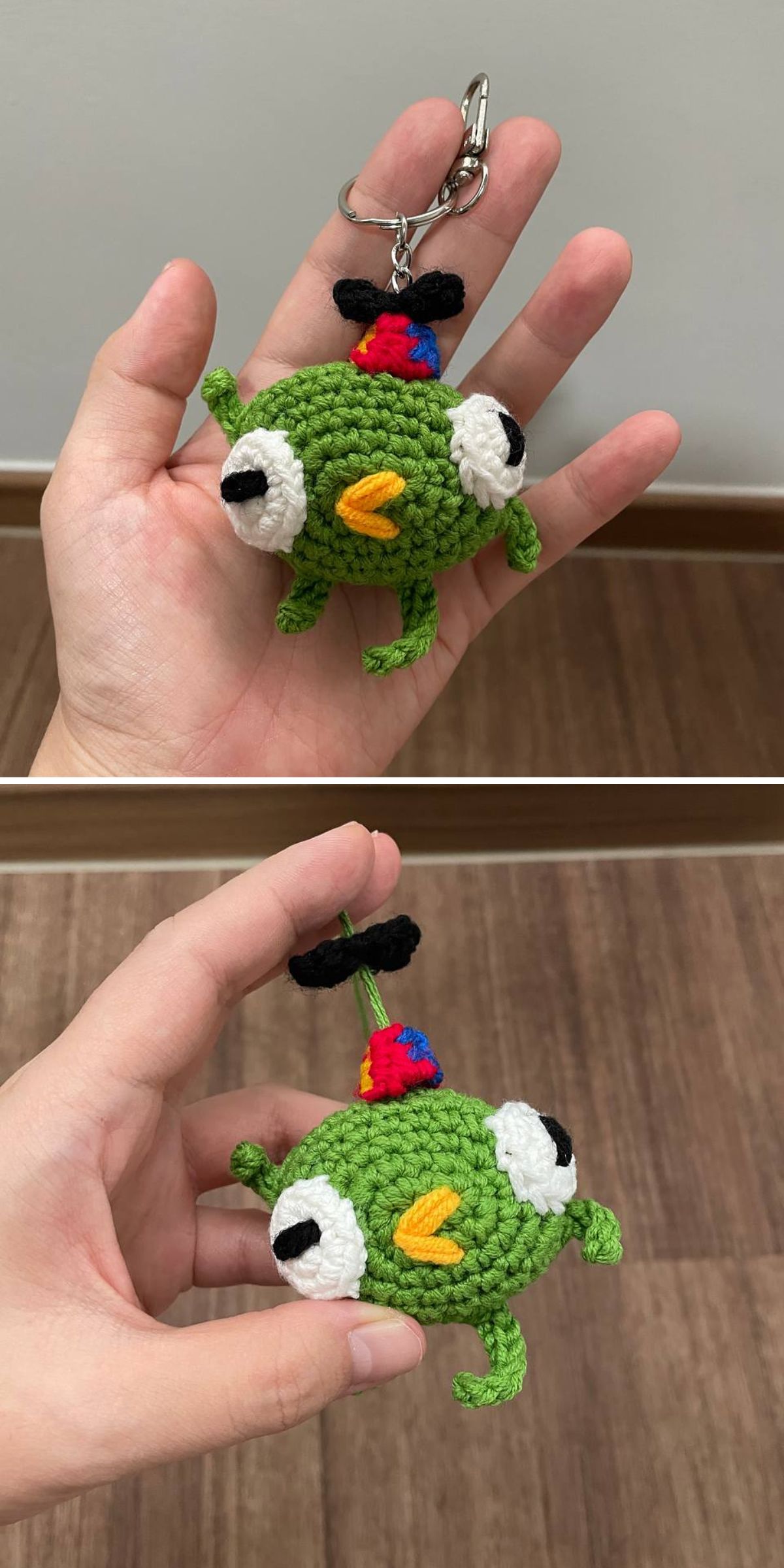 A hand holds a crochet keychain shaped like a green creature, featuring large eyes, a yellow beak, small legs, and a black propeller hat.