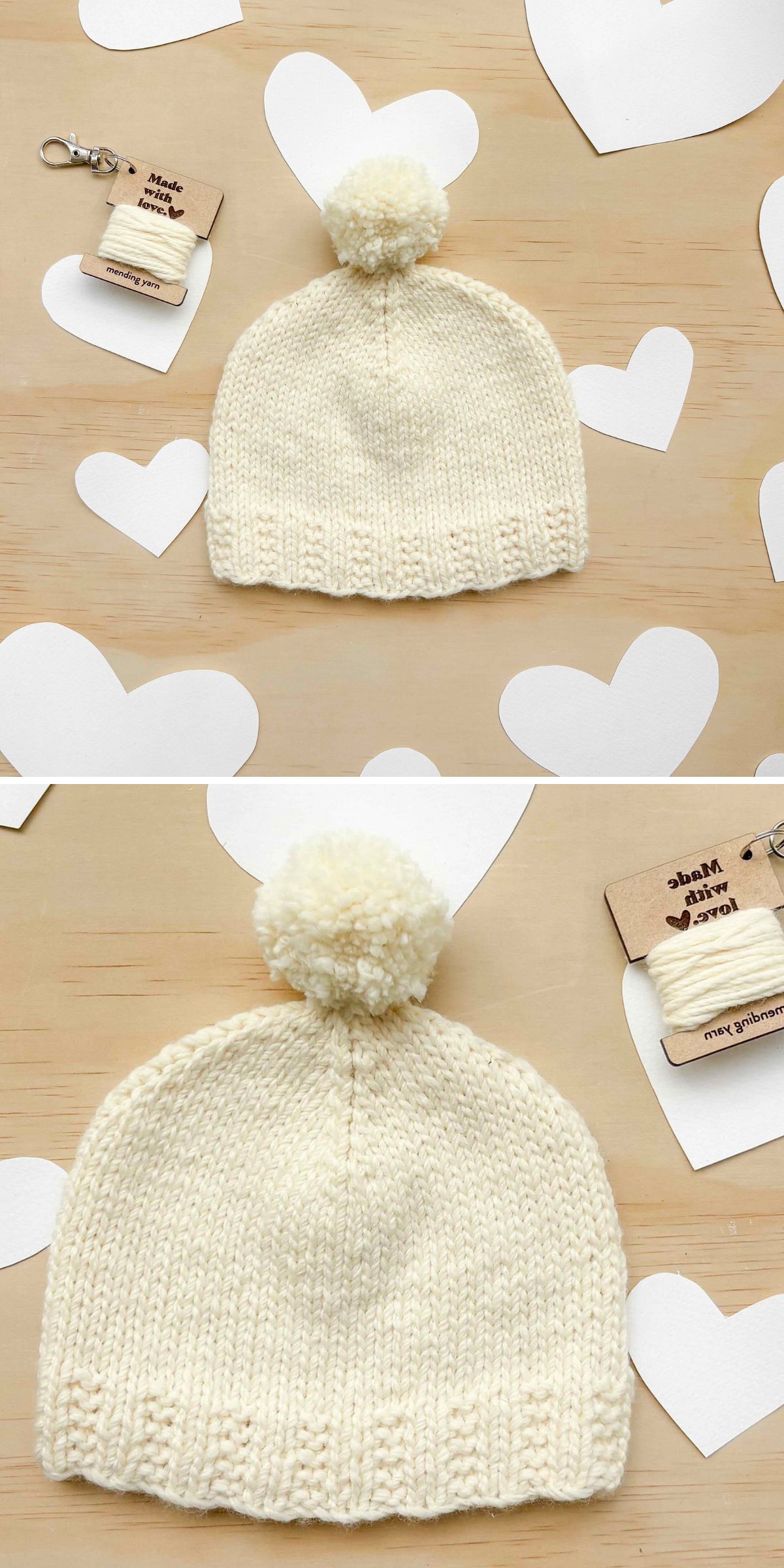 Cream-colored knit baby hat with a pom-pom on top, displayed on a wooden surface with white paper hearts and a product tag nearby.