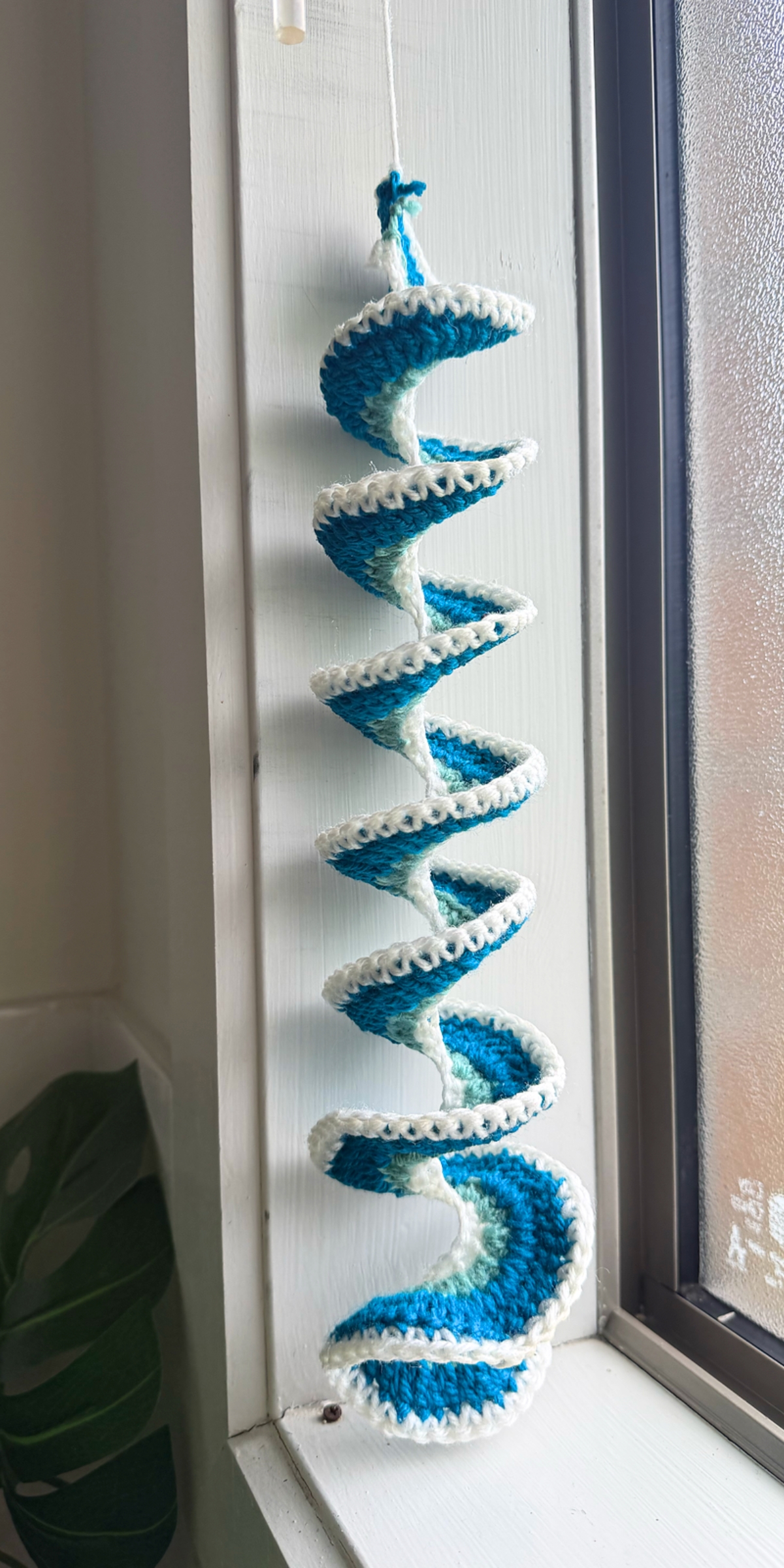 Crocheted blue and white spiral crochet windspinner hanging decoration displayed on a white window frame next to a frosted window.