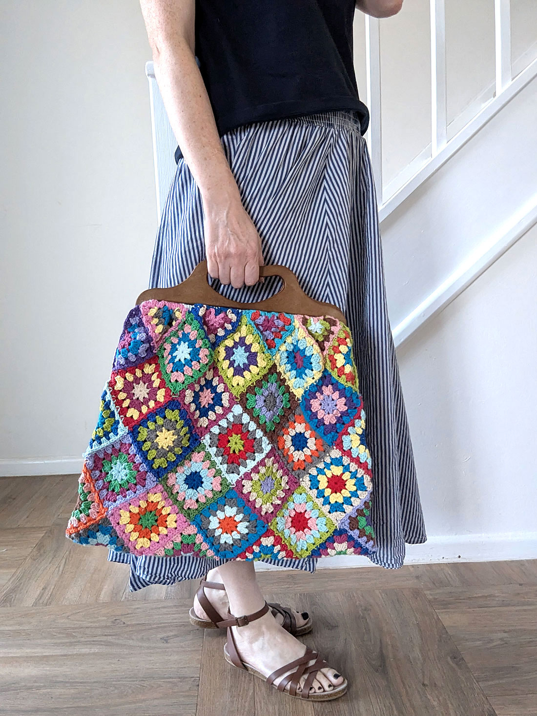 Person holding a colorful granny square bag with wooden handles, standing indoors on a wooden floor, wearing a striped skirt and brown sandals.